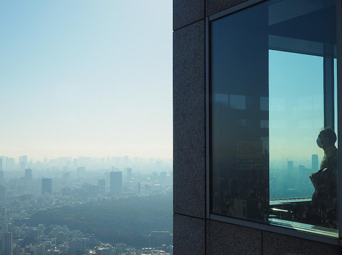 View through a window of the city clouded in smog, a man is standing inside with a mask over his nose and mouth